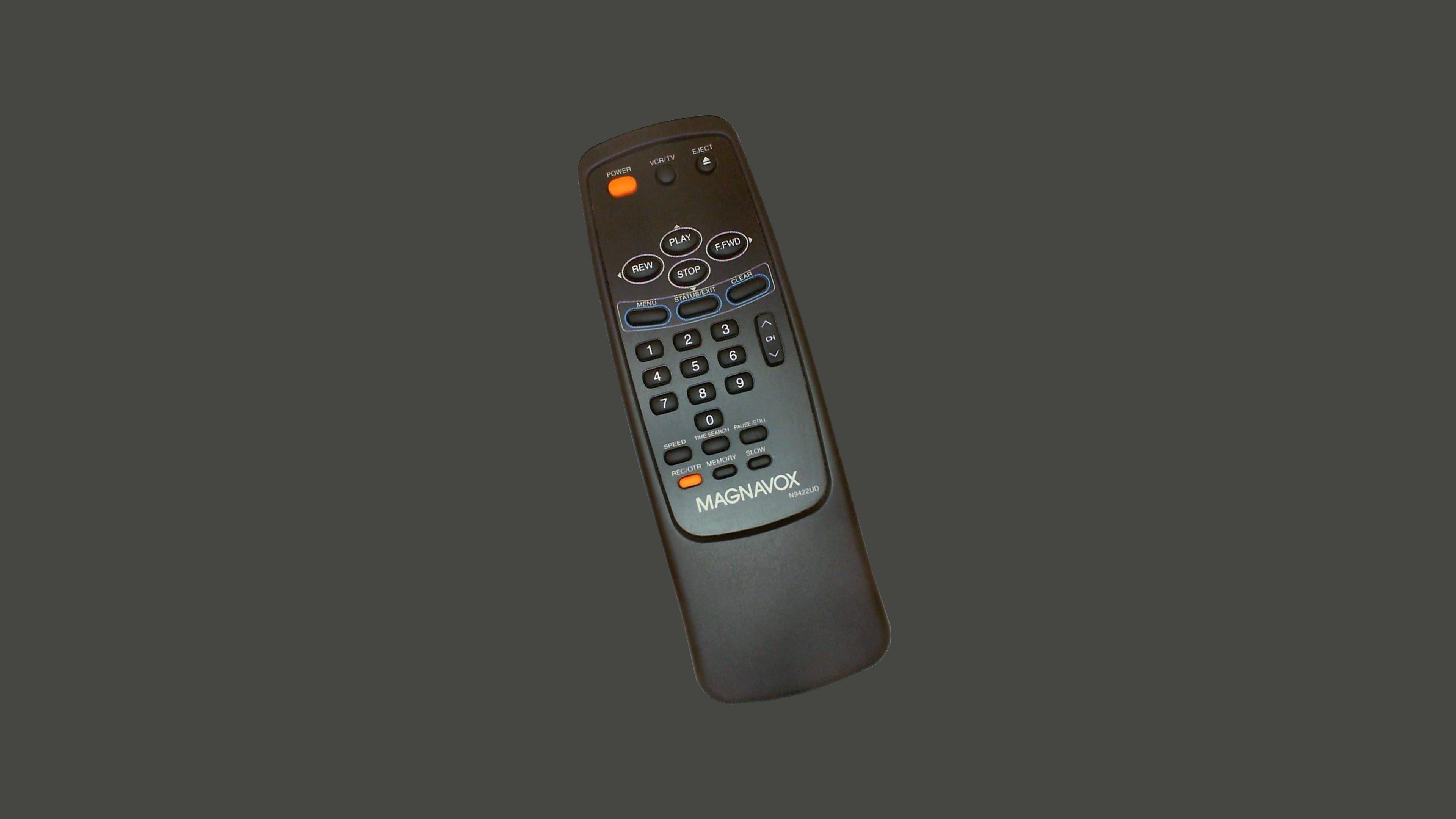 A Magnavox remote control from the 1990s, featuring a black plastic body with a slightly curved shape. At the top, a cluster of playback buttons is arranged in a circular layout, resembling a D-pad, and includes 'Play,' 'Rewind,' 'Fast Forward,' and 'Stop.' Below, there are additional buttons for number input, recording, and other TV functions.
